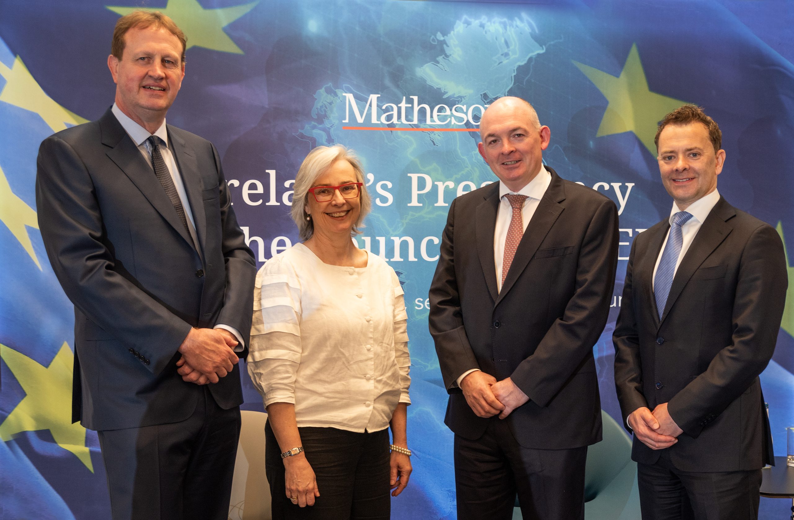 Four people standing side by side in front of a blue backdrop featuring the Matheson logo and a stylised map of Europe with EU stars. The individuals are dressed in business attire, including suits and a light-coloured blouse. The backdrop includes partially visible text referencing Ireland’s presidency and a related event.