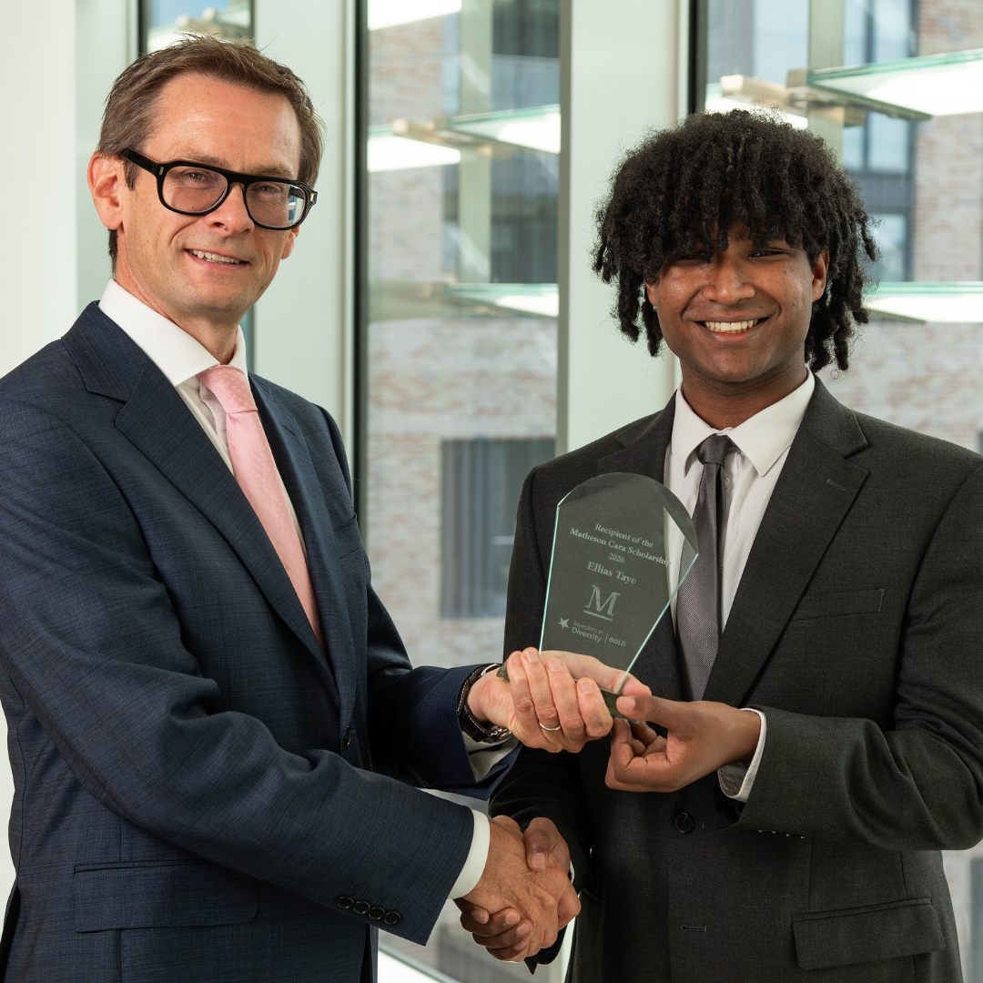 Two people in business attire stand indoors in front of large floor-to-ceiling windows, shaking hands while jointly holding a glass award. The award recipient stands on the right holding the plaque, while the presenter stands on the left. The award contains engraved text recognising the recipient of the 2026 Matheson Cara Scholarship. A modern office building is visible through the windows in the background.