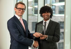 Two people in business attire stand indoors in front of large floor-to-ceiling windows, shaking hands while jointly holding a glass award. The award recipient stands on the right holding the plaque, while the presenter stands on the left. The award contains engraved text recognising the recipient of the 2026 Matheson Cara Scholarship. A modern office building is visible through the windows in the background.