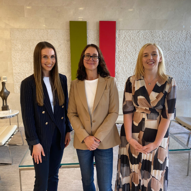 Three people standing side by side in a modern office reception area. The group is positioned in front of a light stone wall featuring two vertical coloured panels. Contemporary white seating, glass tables and a computer screen are visible in the background. All three individuals are facing the camera and posed for a professional photograph.